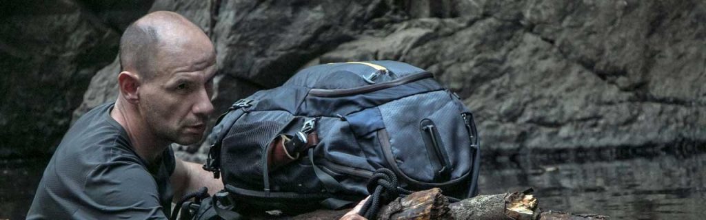 Photo of a survivalist crossing a river protecting his backpack on a floating log.