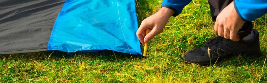 Young man pinning down a waterproof nylon blanket for resting on the grass, with yellow plastic tent stakes or pegs.