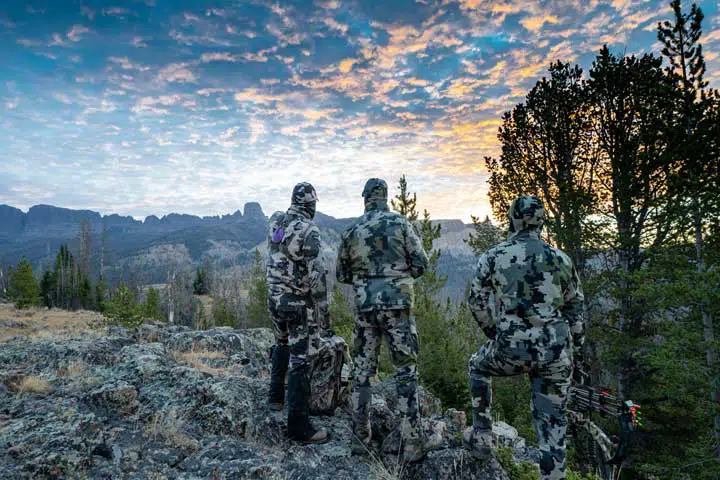 Three adult male hunter friends, unrecognizable, stand on a mountain ridge looking for elk to hunt during bow archery season. 