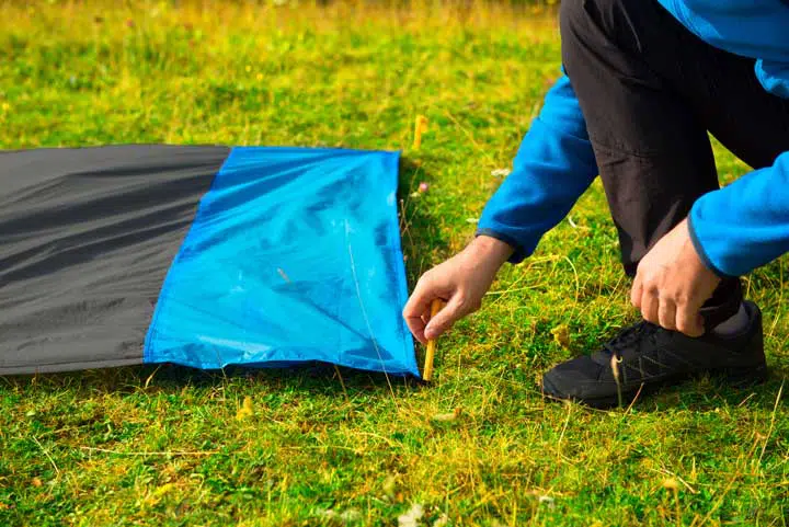 Young man pinning down a waterproof nylon blanket for resting on the grass, with yellow plastic tent stakes or pegs. 