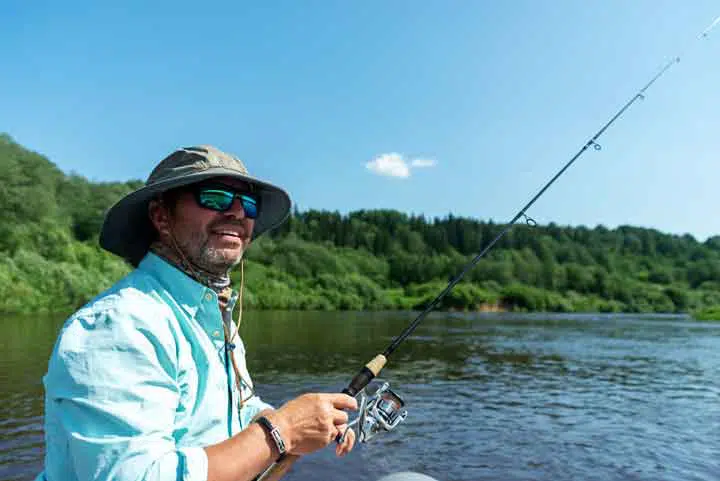 a man wearing a bucket had fishing from a boat on the river with a spinning rod. 