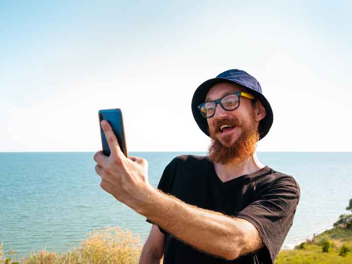 Bearded millennial man in black t-shirt and bucket hat on blue sea landscape smartphone in hand talking taking selfie.