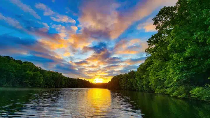 Glow of sunrise on partly clouded day over tree lined lake with reflection. 