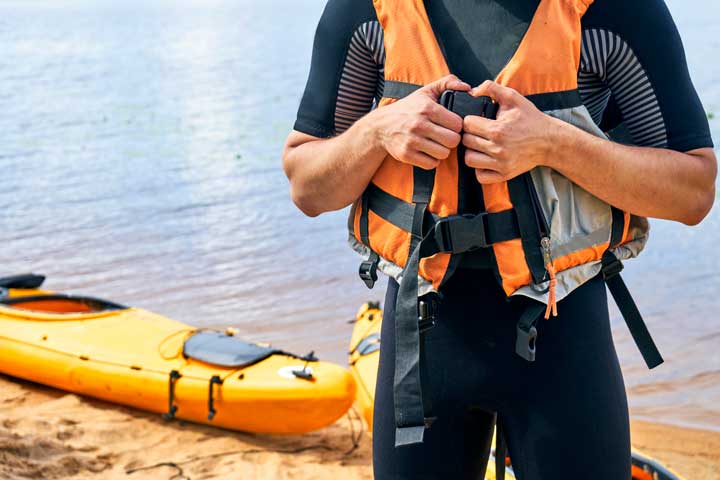 Young male hiker wearing wetsuit putting on a life vest with a yellow kayak. 